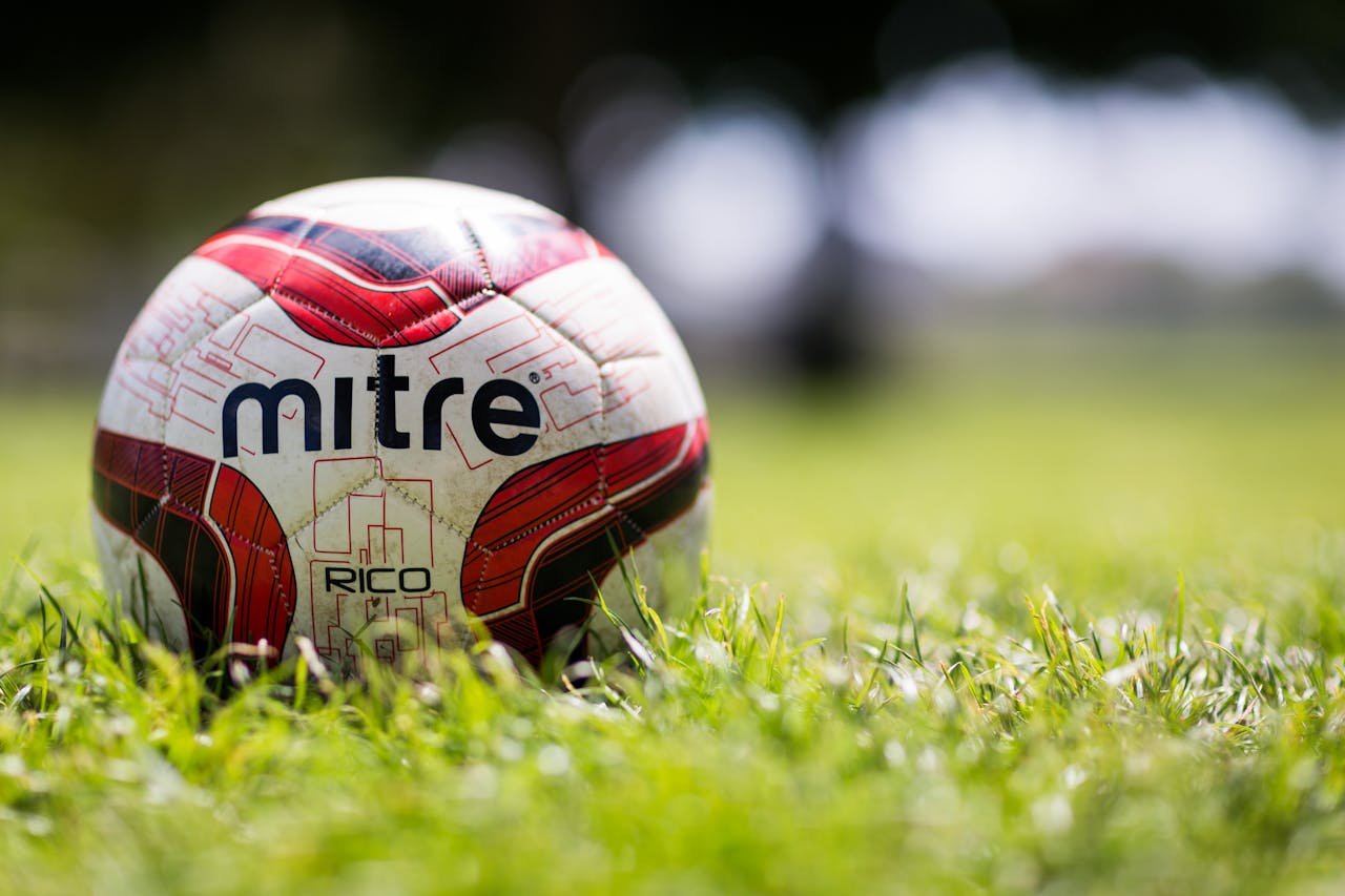 A close-up of a Mitre soccer ball resting on a grassy field with a blurred background.