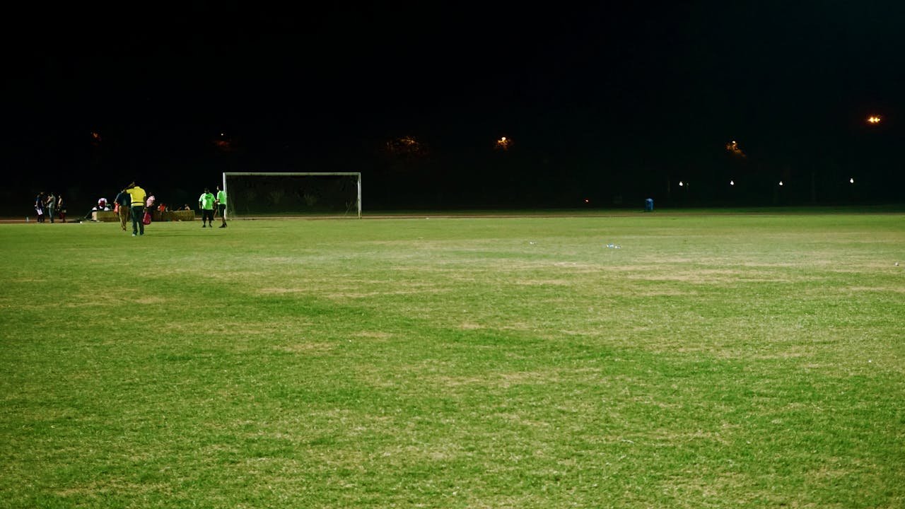 View of a soccer field at night with players and goalposts under floodlights.